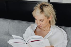 Short blond-haired woman reading a book on a gray couch.